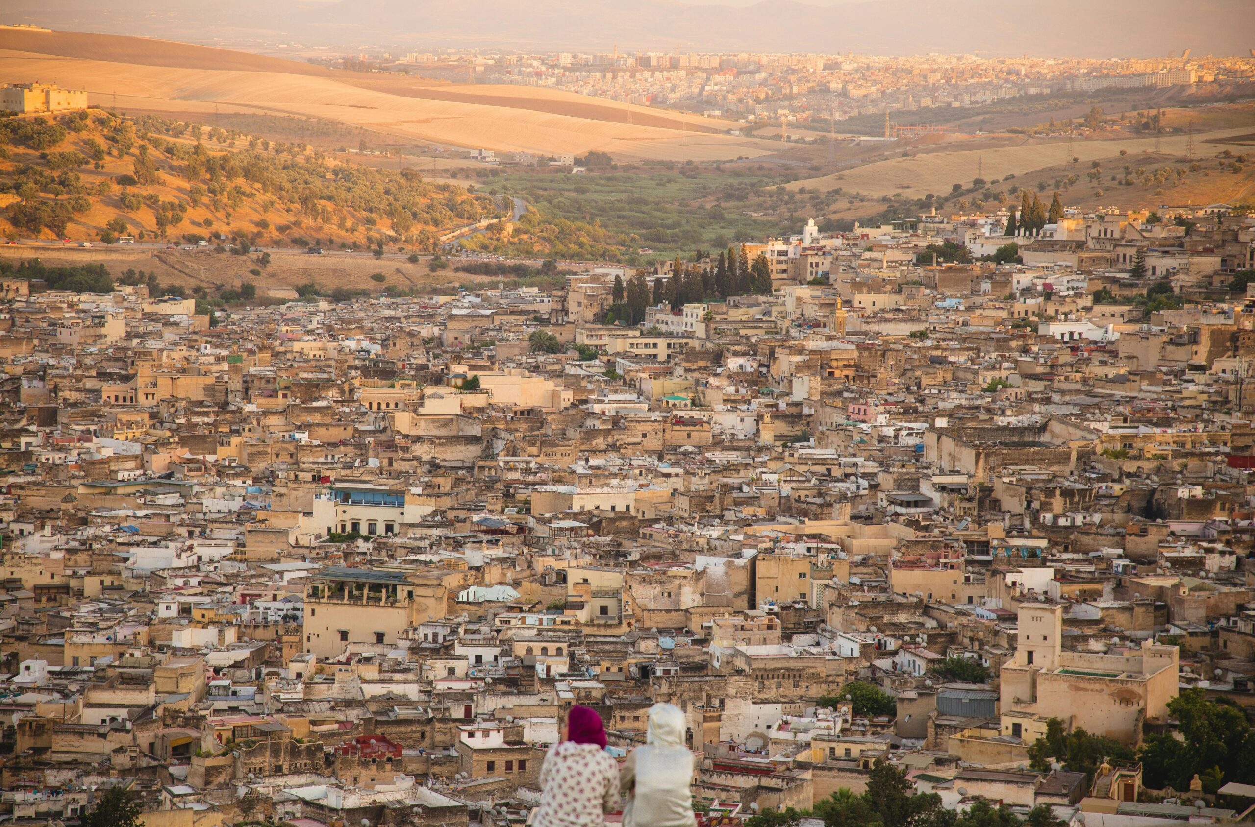 From above back view of unrecognizable female friends in traditional wear sitting on hilltop and looking at town with typical buildings